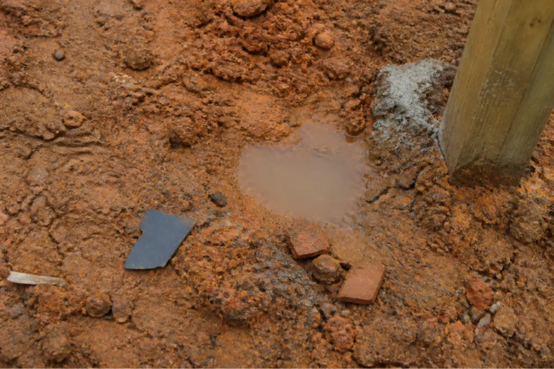 Close-up of compacted orange clay subsoil with brick bits and a puddle that won't drain, typical of a handed-over plot