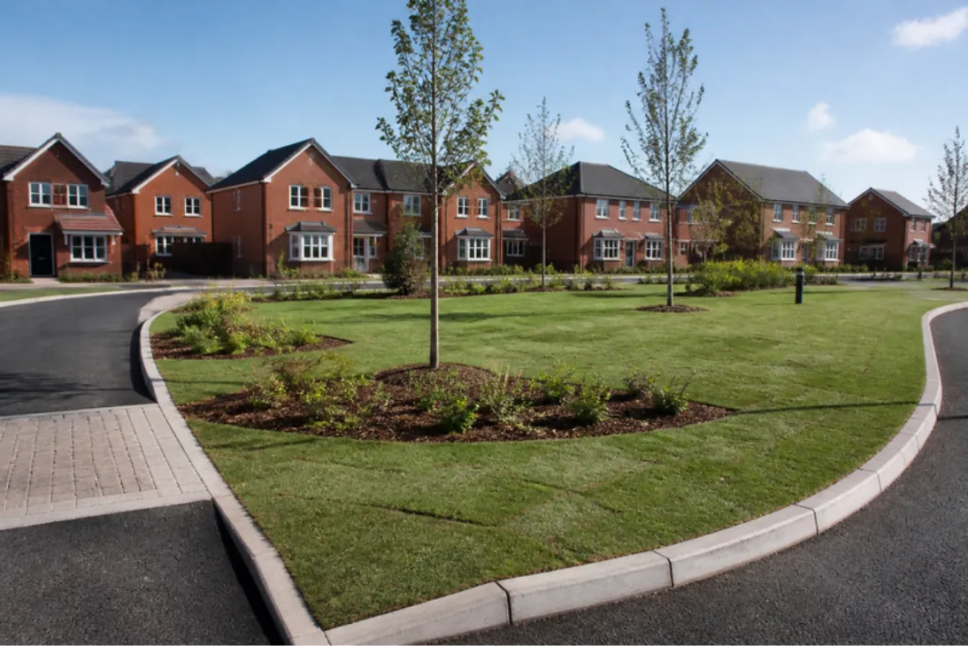 Newly turfed verges and planted island at the entrance to a new housing estate with red-brick homes beyond