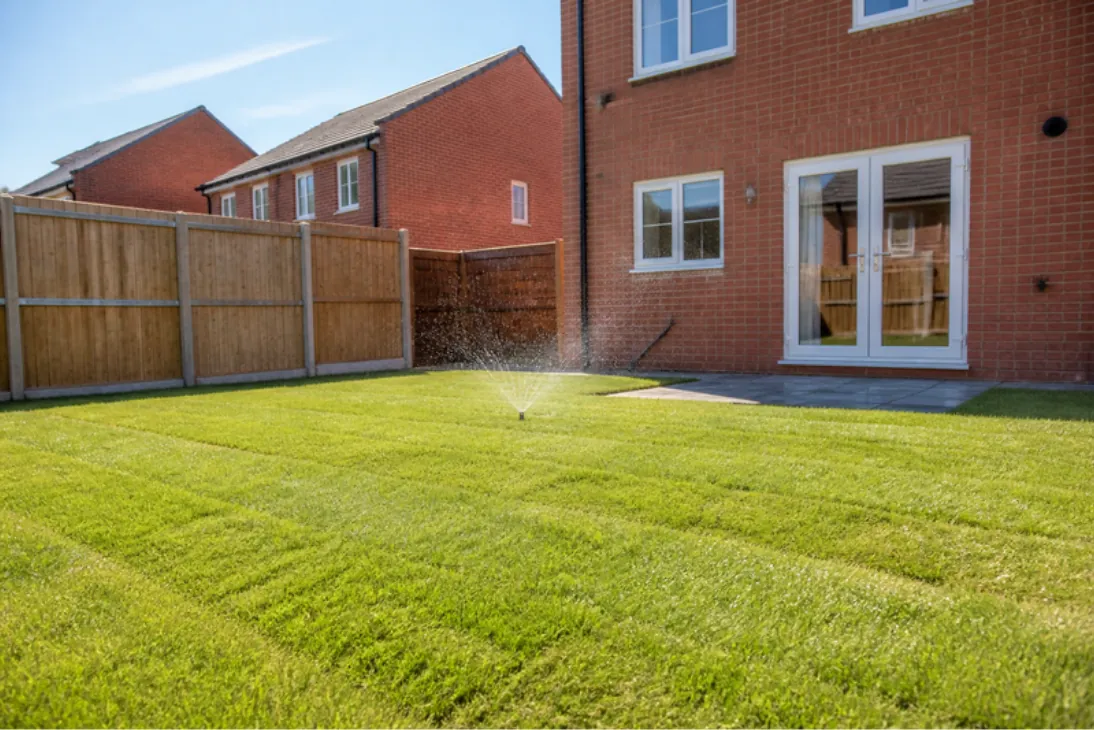 Sprinkler watering a just-laid lawn behind a red-brick new-build semi on a sunny late afternoon