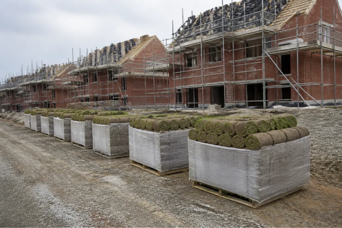 Pallets of rolled turf lined up on a hardcore site road beside part-built red-brick houses and scaffolding